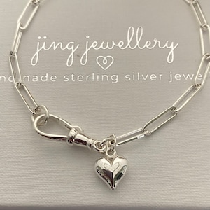 May include: A silver paperclip chain bracelet with a heart charm. The bracelet has a lobster clasp and a small, detailed heart pendant. The background includes the text "jing jewellery" and "handmade sterling silver jewe".