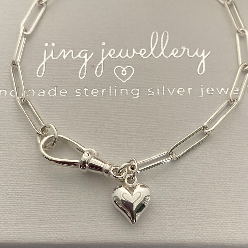 May include: A silver paperclip chain bracelet with a heart charm. The bracelet has a lobster clasp and a small, detailed heart pendant. The background includes the text "jing jewellery" and "handmade sterling silver jewe".