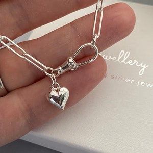May include: A silver necklace with a heart charm. The necklace has a paperclip chain and a lobster clasp. The heart pendant is small and polished, reflecting light. The jewellery is likely made of sterling silver. The image shows the word 'jewellery'.