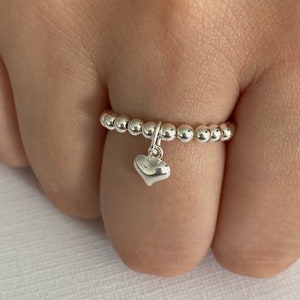 May include: A silver beaded ring with a dangling heart charm. The ring is made of small, round beads and features a polished heart pendant. The ring is shown on a finger.