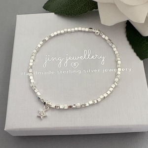 May include: A silver bracelet featuring square beads and a star pendant, displayed on a white box. The box is printed with "jing jewellery" and "Handmade sterling silver jewellery". A delicate piece of jewellery.