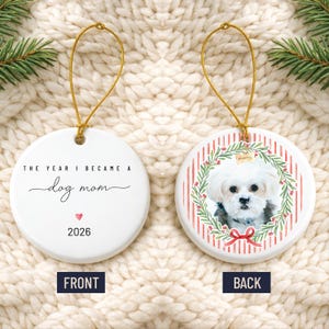 Op de afbeelding: Een rond wit keramieken ornament met een gouden ophangkoord. De voorzijde bevat de zwarte tekst THE YEAR I BECAME A dog mom, een klein rood hartje en 2026. De achterzijde toont een foto van een witte hond in een groene krans met rode bessen.