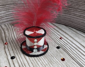 Può includere: Un piccolo cappello a cilindro rosso e bianco con una fascia nera e una piuma rossa. Il cappello è decorato con glitter argentato e strass.