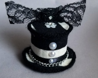 Può includere: Un cappello a cilindro a righe bianche e nere con un fiocco di pizzo nero e decorazioni di perle. Il cappello è decorato con una catena argentata.