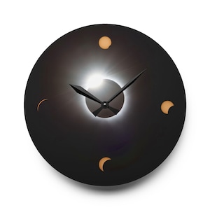 Puede incluir: Un reloj de pared redondo negro con un diseño de eclipse solar. La esfera del reloj presenta un centro blanco brillante, manecillas negras y cuatro pequeñas formas de media luna y circulares iluminadas. El reloj está sobre un fondo blanco.