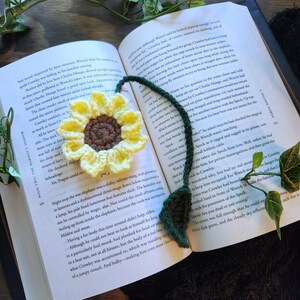 Könnte beinhalten: Gehäkelter Sonnenblumen-Lesezeichen mit gelben Blütenblättern und brauner Mitte, auf den Seiten eines Buches liegend. Das Lesezeichen hat einen grünen Stiel und ein Blatt.