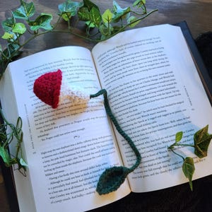 Könnte beinhalten: Handgefertigter Häkel-Lesezeichen in Pilzform mit rotem Hut und weißem Stiel sowie einem grünen Blatt. Das Lesezeichen befindet sich in einem offenen Buch. Ein einzigartiges Accessoire für Bücherliebhaber.