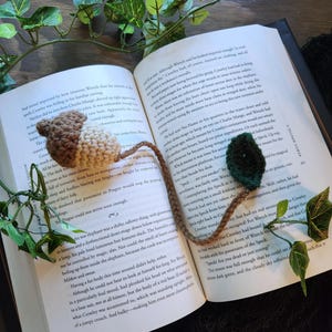 Könnte beinhalten: Handgefertigter Häkel-Lesezeichen mit einem braunen und beigen Eicheldesign, einem langen braunen Stiel und einem grünen Blatt. Das Lesezeichen liegt auf den offenen Seiten eines Buches, mit dekorativem Efeu.