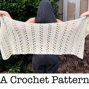 May include: Cream-colored crochet shawl with a textured, openwork design. The shawl is held up, showcasing its rectangular shape and intricate stitch pattern. The text "A Crochet Pattern" is displayed at the bottom of the image.