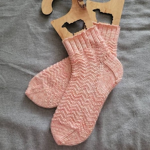 May include: A pair of pink knitted socks with a chevron pattern. The socks are displayed on a wooden hanger with sheep silhouettes.