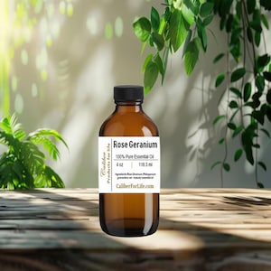 May include: A brown glass bottle with a black cap, labeled "Rose Geranium 100% Pure Essential Oil, 4 oz, 118.3 ml." The bottle is set against a blurred background of greenery and a wooden surface. The label also includes the text "Caliber Products for life" and "CaliberForLife.com."