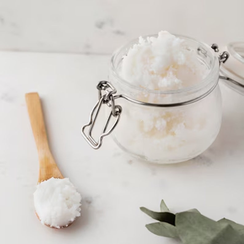 May include: A clear glass jar with a metal clasp filled with white, textured cream. A wooden spoon holds a scoop of the cream. The items are on a white surface, suggesting a skincare or cosmetic product.