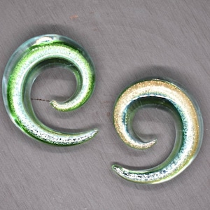 May include: A pair of green and gold spiral glass ear gauges. The gauges have a silver glitter finish.
