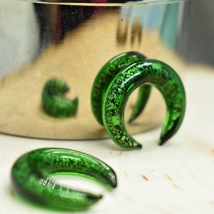 May include: Three green glass ear gauges with a speckled finish. The gauges are shaped like a crescent moon.