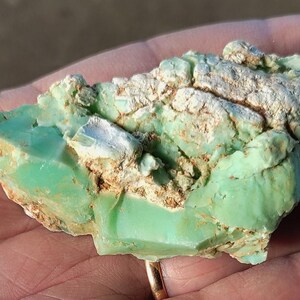 Può includere: Una gemma naturale, tagliata in modo grezzo, che mostra un vivace colore verde chiaro. La pietra ha una superficie strutturata con aree bianche e beige. La gemma è tenuta in una mano, evidenziando la sua forma irregolare e la sua bellezza naturale.