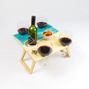 May include: A wooden picnic table with a blue and white painted top. The table has built-in wine glass holders and a small bowl for snacks. Four wine glasses and a bottle of wine are on the table.