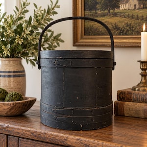 May include: A cylindrical black wooden bucket with a matching lid and a tall, arched handle. The container features visible horizontal bands secured with small metal fasteners, showcasing a weathered, matte finish on its exterior surface.