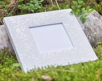 Puede incluir: Un marco de fotos blanco con un patrón decorativo se encuentra sobre un lecho de musgo verde. El marco tiene un paspartú blanco y una ventana transparente para una foto.