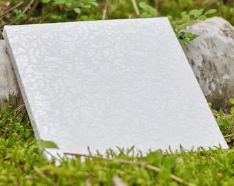 Puede incluir: Un lienzo blanco con un delicado patrón de encaje, descansando sobre un lecho de musgo verde. El lienzo está ligeramente inclinado, creando una sensación de profundidad.