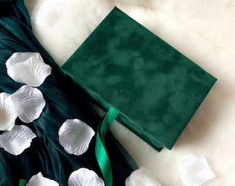 May include: A dark green velvet box with a green ribbon on a white fluffy surface. White flower petals are scattered around the box.