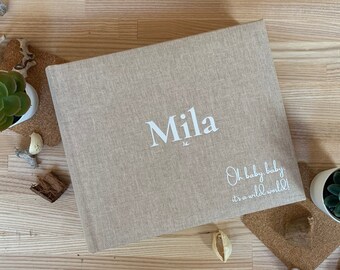May include: A beige linen-covered photo album with the name "Mila" in white script. The text "Oh baby, baby it's a wild world!" is below. The album is on a wooden surface, with small potted succulents and cork coasters.