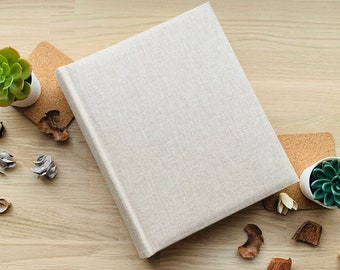 May include: A beige linen-covered photo album with a textured surface. The album is lying on a light wood surface with small decorative items and potted succulents.