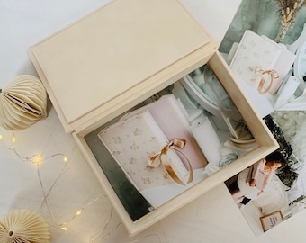 May include: A white wooden box with a hinged lid, open to reveal a stack of books with floral patterned covers and a gold ribbon. The box is sitting on a white surface with a string of lights and paper ornaments.