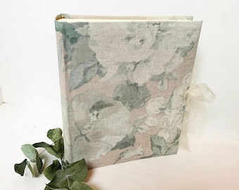 May include: A floral patterned linen-covered scrapbook with a light pink and green color scheme. The scrapbook has a ribbon tied around it.