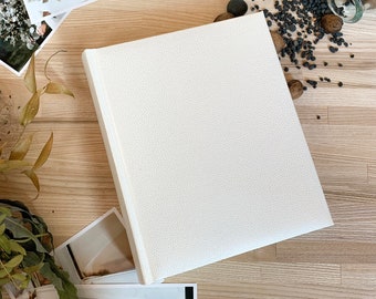 May include: A white leather photo album with a textured surface. The album is closed and lying flat on a wooden surface.
