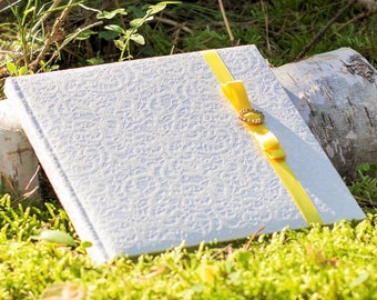 Puede incluir: Un diario blanco de tapa dura, estampado, con una cinta amarilla atada alrededor. El diario está sobre un lecho de musgo verde.