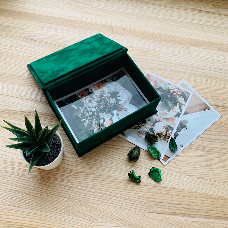 May include: A dark green velvet photo box with a hinged lid, open to reveal a photograph. Several printed photos and a small potted plant are next to the box. Dried green leaves are scattered around the photos.