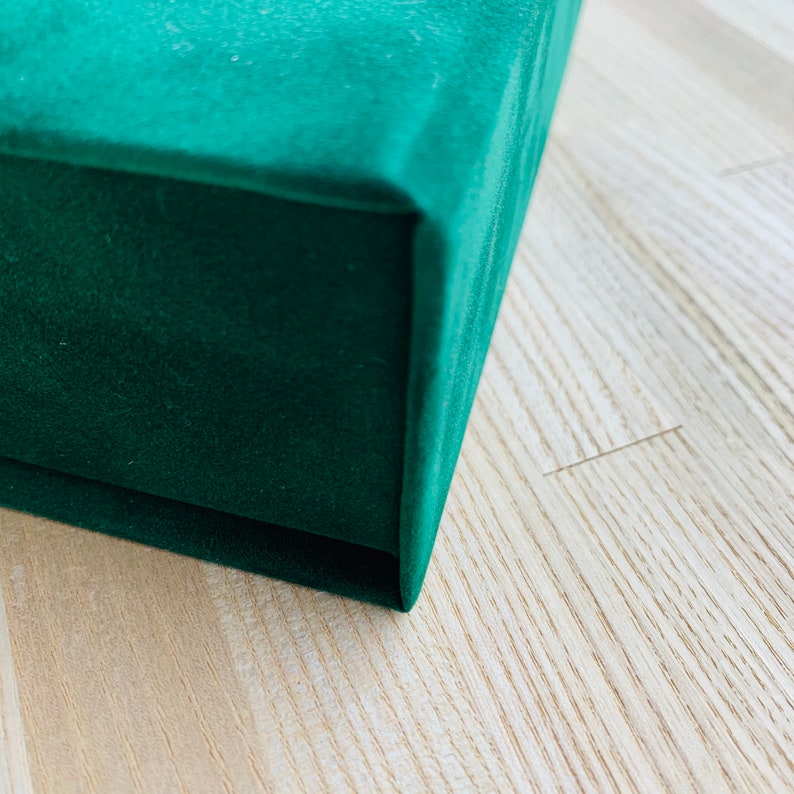 May include: Close-up of a dark green velvet box. The box has a soft, plush texture and a deep, rich colour. The box is sitting on a light-coloured wooden surface.