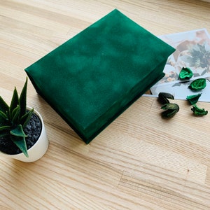 May include: A rectangular, emerald green velvet box with a matching lid. The box is closed and sits on a light wood surface, next to a small potted succulent and a decorative print with dried botanicals. The box is a storage container.