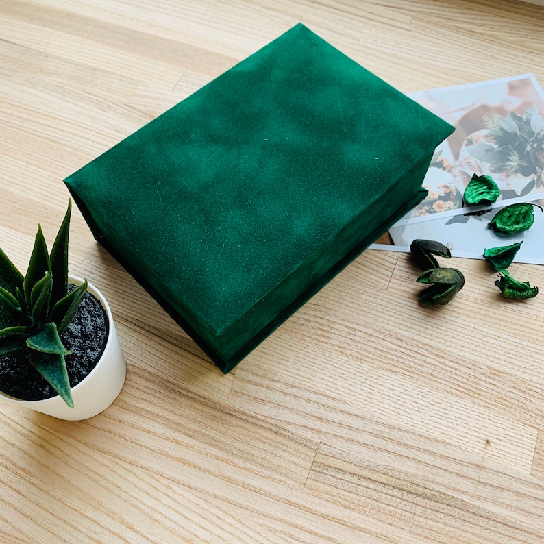 May include: A rectangular, emerald green velvet box with a matching lid. The box is closed and sits on a light wood surface, next to a small potted succulent and a decorative print with dried botanicals. The box is a storage container.