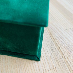 May include: Close-up of a dark green velvet box. The box is rectangular with a lid. The velvet material has a soft, plush texture. The box is on a light-coloured wooden surface.