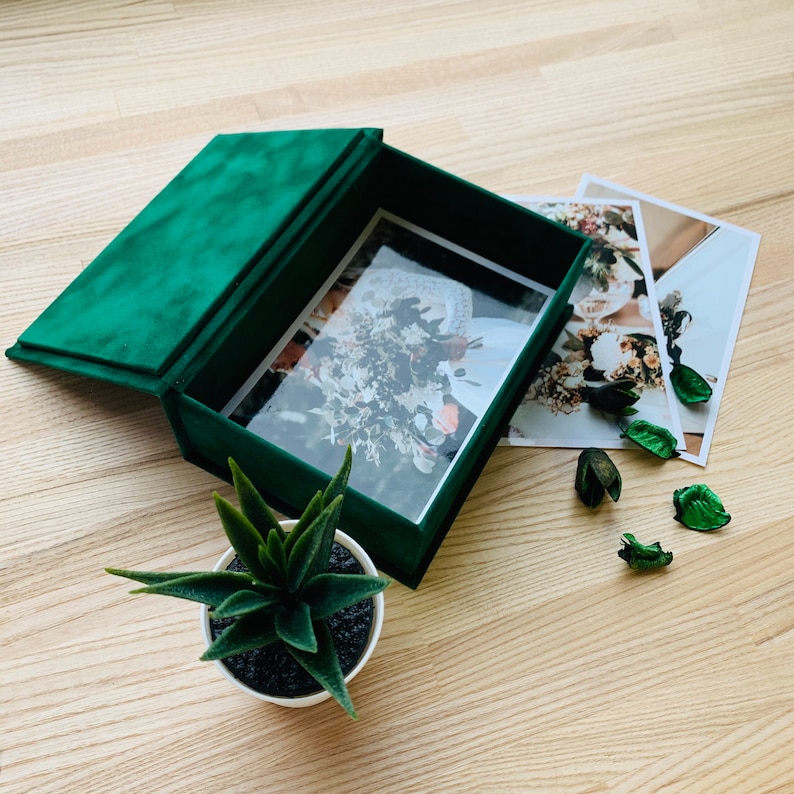 May include: A dark green velvet photo box with a hinged lid, open to reveal a photograph. Two additional photos are next to the box. A small succulent plant in a white pot and decorative green potpourri are also present.