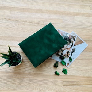 May include: A dark green velvet photo album with printed photographs of floral arrangements. The album is open, next to a small potted succulent and decorative dried leaves. The background is a light wood grain. The album is rectangular.