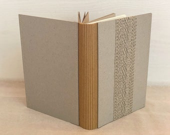 May include: A closed book with a brown cardboard cover and a spine with a decorative brown paper strip. The front cover has a decorative lace trim.