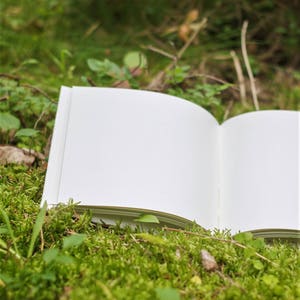 Puede incluir: Un libro abierto en blanco que descansa sobre un lecho de musgo verde.