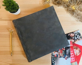 Puede incluir: Un álbum de fotos de tapa blanda de color gris oscuro con una superficie texturizada. El álbum está sobre una superficie de madera clara con una pequeña planta en maceta y un bolígrafo dorado cerca. Hay varias tarjetas navideñas con fotos de personas y un árbol de Navidad en el fondo.
