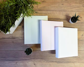 May include: Three blank photo albums, one light green, one light pink, and one white, are arranged on a wooden surface.