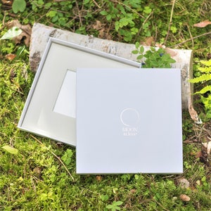 Puede incluir: Una caja blanca con el texto "MOON in love" y un dise&ntilde;o circular. La caja est&aacute; abierta y se ve un marco blanco en el interior. La caja est&aacute; sobre un lecho de musgo verde.
