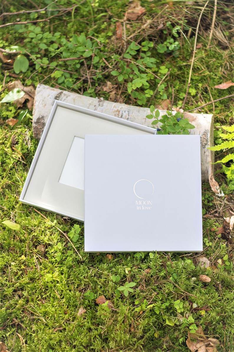Puede incluir: Una caja blanca con el texto "MOON in love" y un dise&ntilde;o circular. La caja est&aacute; abierta y se ve un marco blanco en el interior. La caja est&aacute; sobre un lecho de musgo verde.