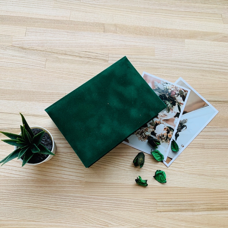 May include: A dark green velvet photo album with printed photographs of floral arrangements. The album is open, next to a small potted succulent and decorative dried leaves. The background is a light wood grain. The album is rectangular.