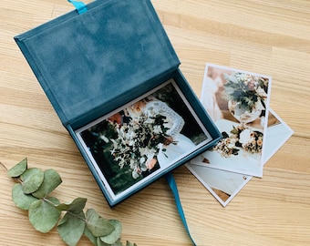 May include: A blue velvet box with a ribbon closure, open to reveal a photograph of a couple holding a bouquet of flowers. The box is on a wooden surface with two additional photographs.