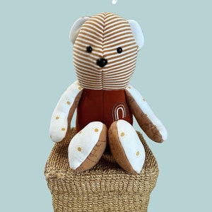 May include: A brown and white striped memory bear sitting on a woven basket. The bear has a brown nose and black eyes. The bear is wearing a red shirt with a rainbow design.