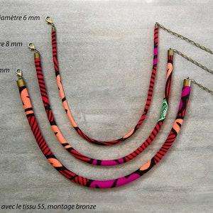 Peut inclure: Trois colliers en tissu de couleurs rouge, rose et orange avec des motifs noirs et blancs. Les colliers ont un diamètre de 6 mm, 8 mm et 10 mm. Chaque collier est doté d'un fermoir en bronze et d'une chaîne en bronze avec une perle rouge à l'extrémité. "Modèles réalisés avec le tissu 55, montage bronze" est écrit en bas de l'image.