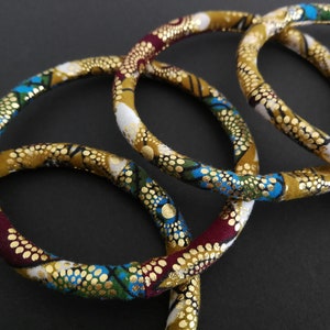 Può includere: Quattro braccialetti in tessuto con un design a motivi dorati, blu, verdi e rossi. I braccialetti sono impilati uno sopra l'altro.