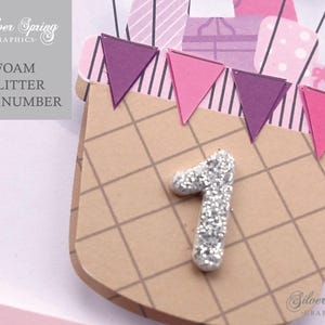 May include: A brown and white checkered paper craft with a silver glitter number one. The craft is decorated with purple, pink, and white paper flags.