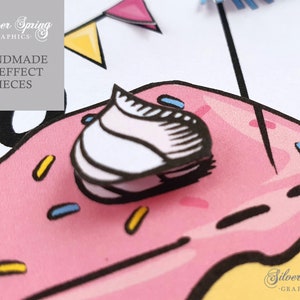 May include: A pink donut with yellow and blue sprinkles and a white frosting swirl on top. The donut is surrounded by a white background with a black outline. The text 'Silver Spring Graphics' is visible in the lower right corner of the image. The text 'Handmade 3D Effect Pieces' is visible in the upper left corner of the image.
